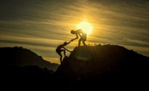 A silhouette of one person reaching his hand out to help another person up a mountain with the sun behind them.