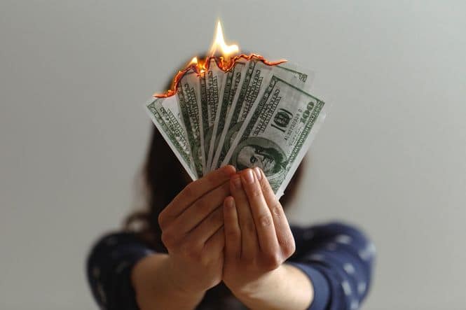 A person holding one hundred dollar bills in front of their face as the bills burn on fire.