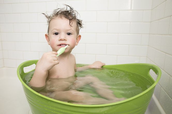 An image of a young child sitting in a tub of bath water while brushing his teeth.