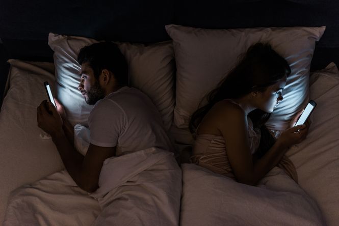 A young married couple showing one of their toxic marriage habits by ignoring the needs of each other while looking at their phones in bed.