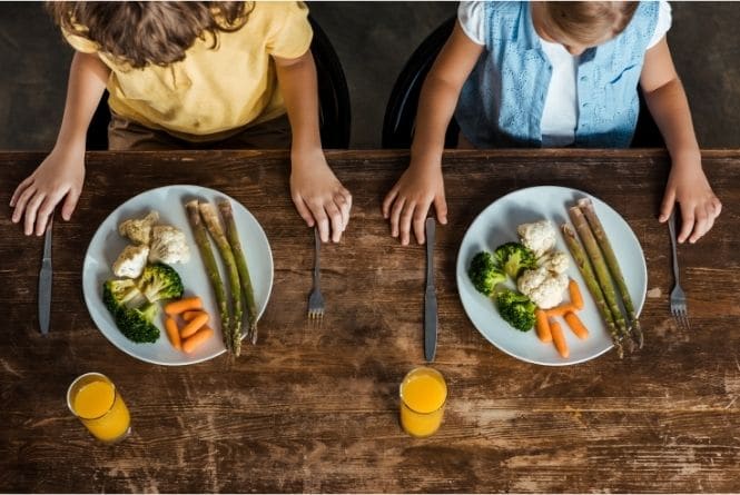 Benefits of having more than one child includes helping each other be healthier. This is a photo from the top down showing two kids sitting at a table with a plate of vegetables in front of them.