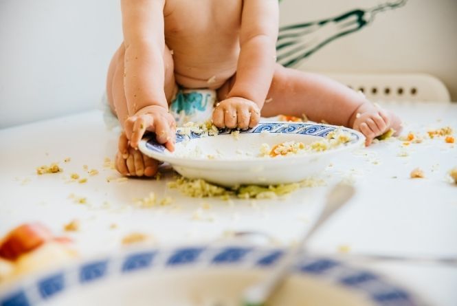 Growing your family does not need to be messy. This is an image of a baby from the chest down sitting on a table making a mess with his food.