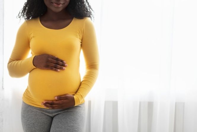 Growing your family will change your body. This image shows a woman from the nose down in a yellow shirt holding her pregnant belly in front of a white background.